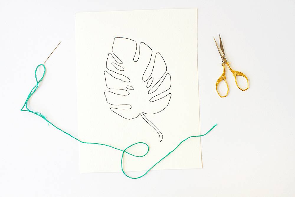 A needle threaded with green thread laying next to a pair of scissors and a piece of paper with a hand drawn leaf on it.