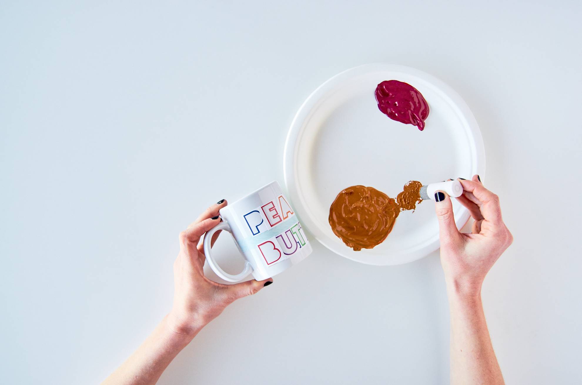 Woman holding a mug in one hand while placing craft paint onto a paper plate with her other hand.