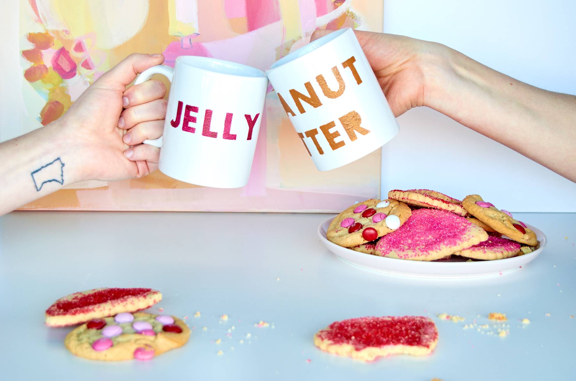 Two women cheers using their decorative mugs over a plate of sugar cookies.