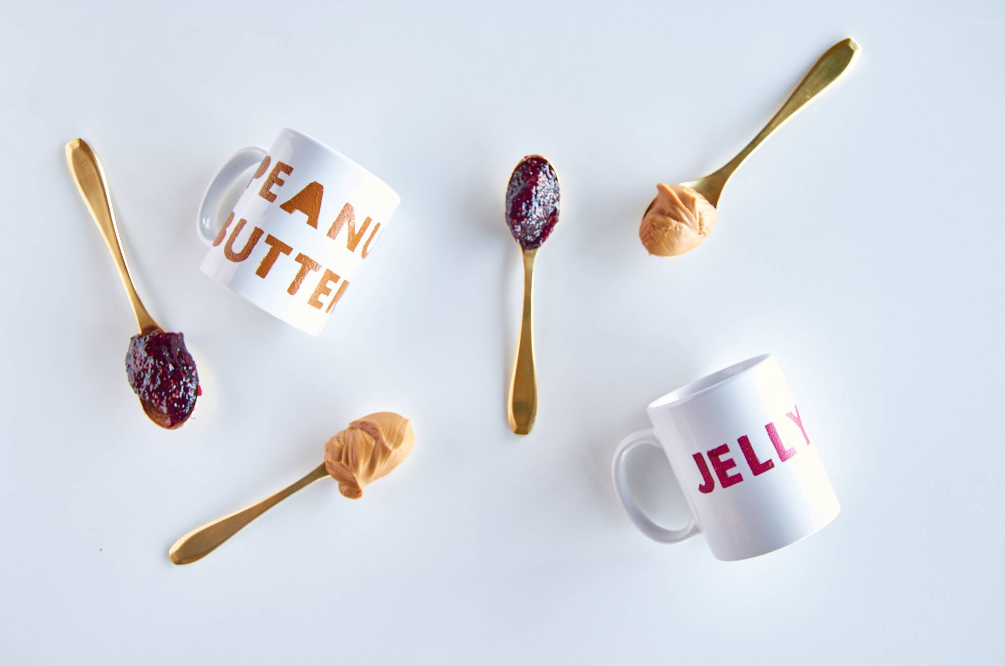 Two spoons with jelly and two spoons with peanut butter scattered about a mug that says peanut butter and a mug that says jelly.