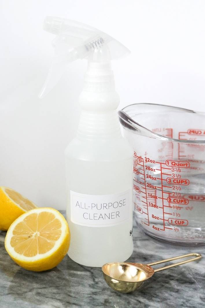 A spray bottle, measuring cup, measuring spoon and two half lemons on a marble counter.