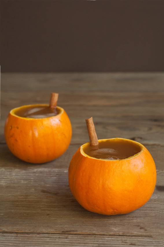 Pumpkins with tops removed containing a drink and cinnamon sticks on top of wood plank table.