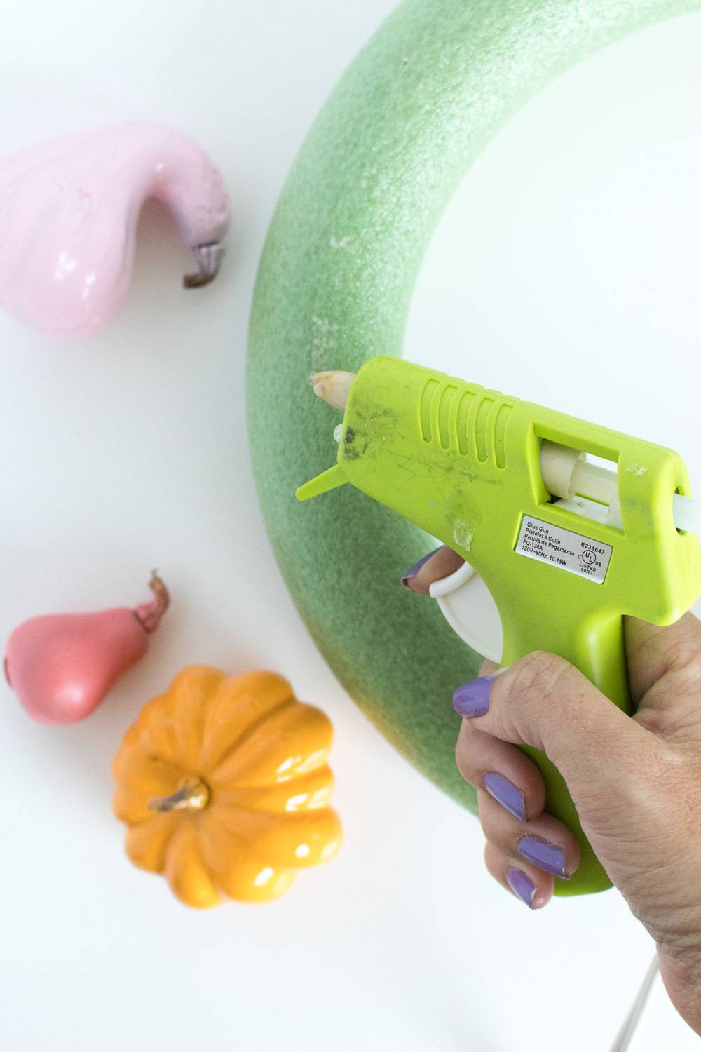 A person is using a hot glue gun to put glue on a green foam ring and there is a small pumpkin and other things near it.