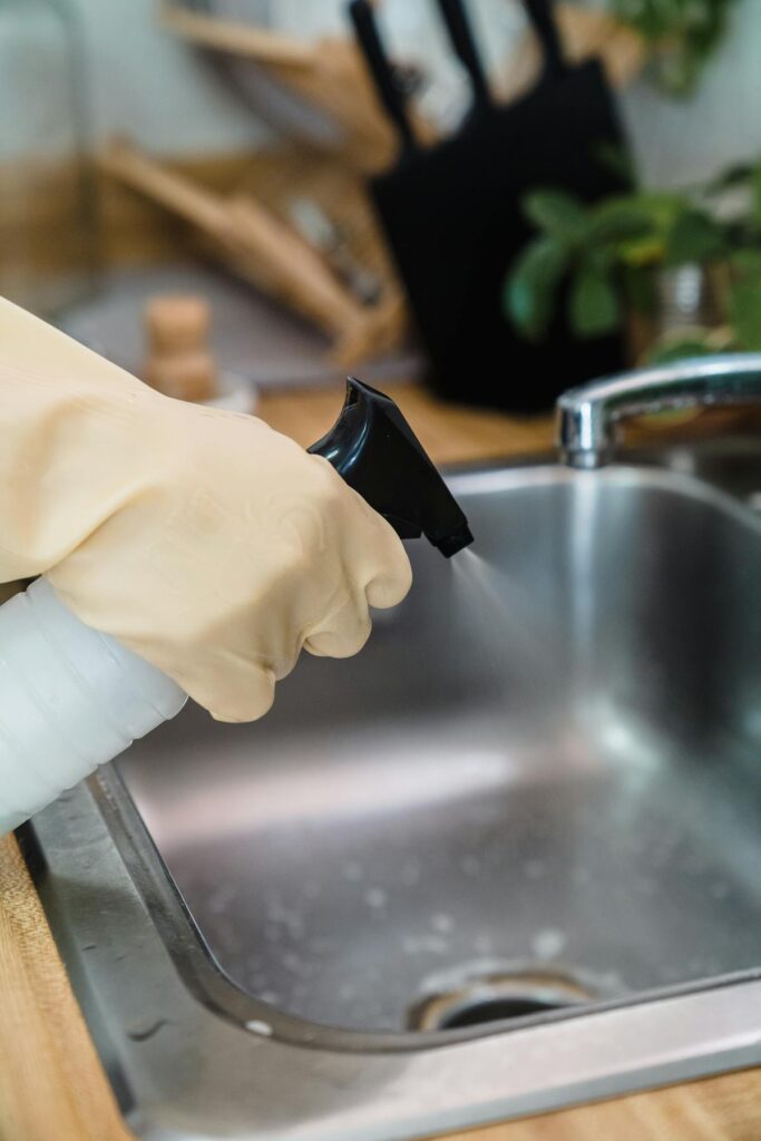 Close-up of a gloved hand cleaning a kitchen sink using a spray bottle, showcasing hygiene.